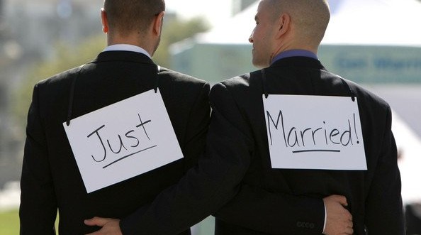 Two grooms facing away from the camera, with "Just" and "Married" signs taped to their backs.