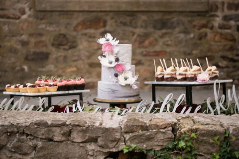 White wedding cake with white and pink flowers, cupcake platters on the side, in front of a stone wall.