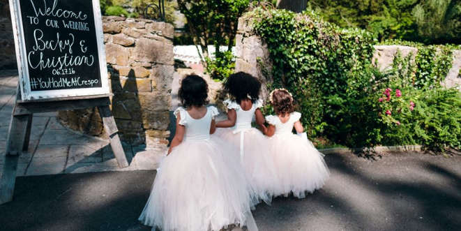 Three young girls turned around, in white dresses with pink skirts near a stone wall.