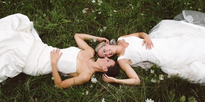 Two brides laying face-up on the grass, looking at each other lovingly.