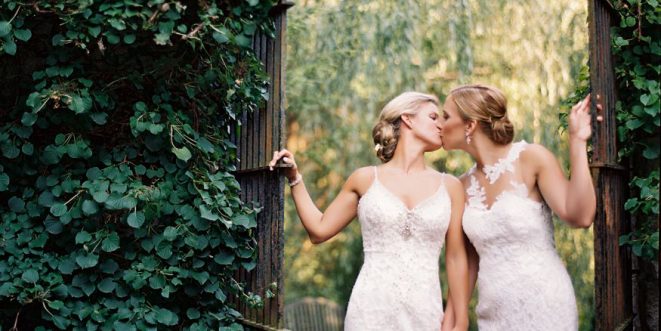 Two brides sharing a kiss under a leaf-ridden archway.