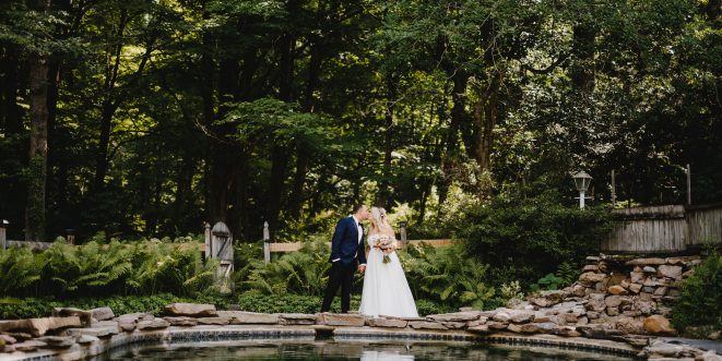 Bride and groom sharing a kiss in front of a pond, with a forest of green trees in the background.