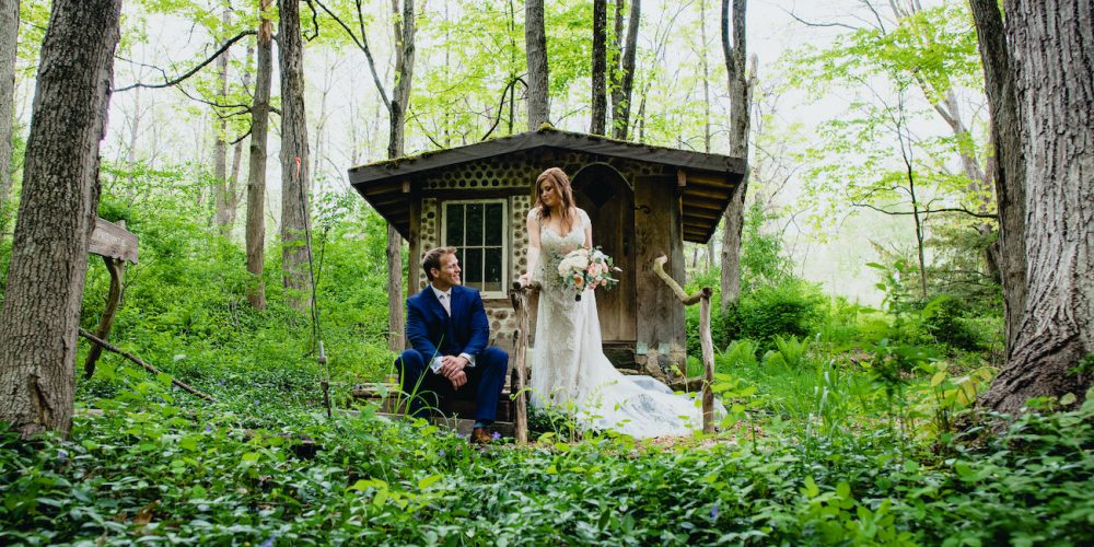 Bridge and groom pose in front of wooden structure in the woods, groom sitting, bridge standing and holding bouquet