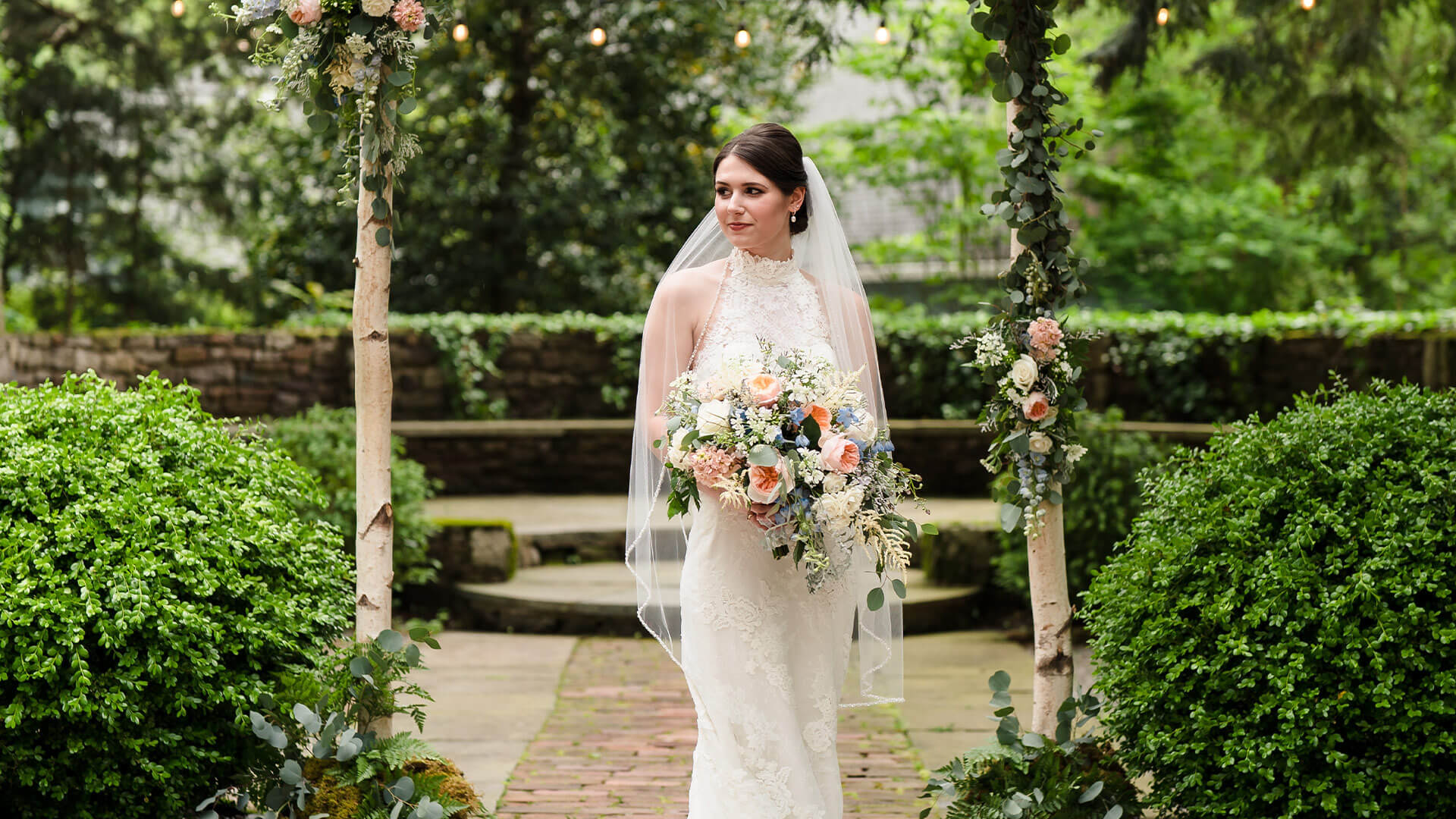 Bride standing under a wooden archway in a courtyard holding her bouquet.
