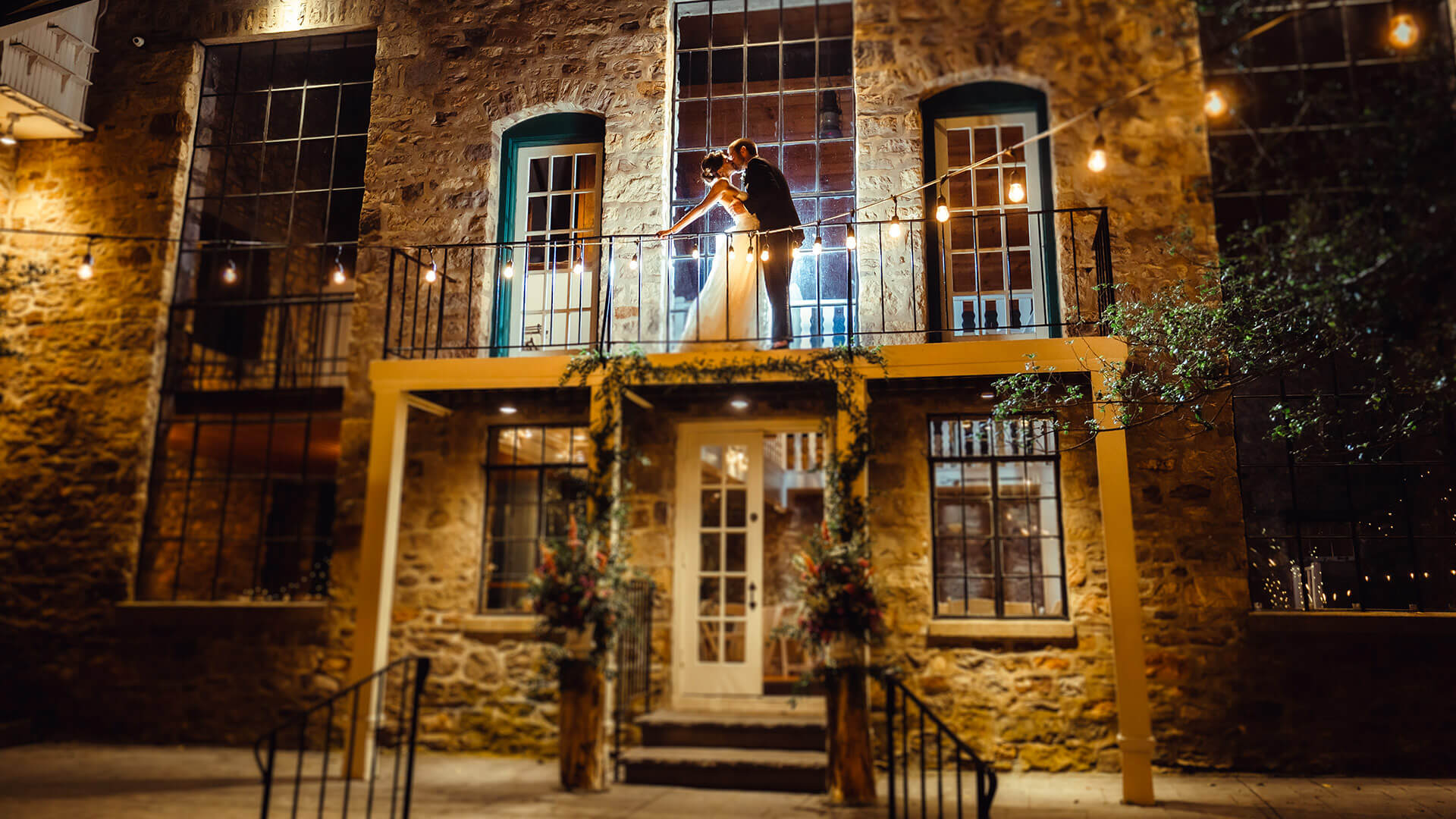 Bride and groom sharing a passionate kiss on a stone building’s illuminated balcony at night.