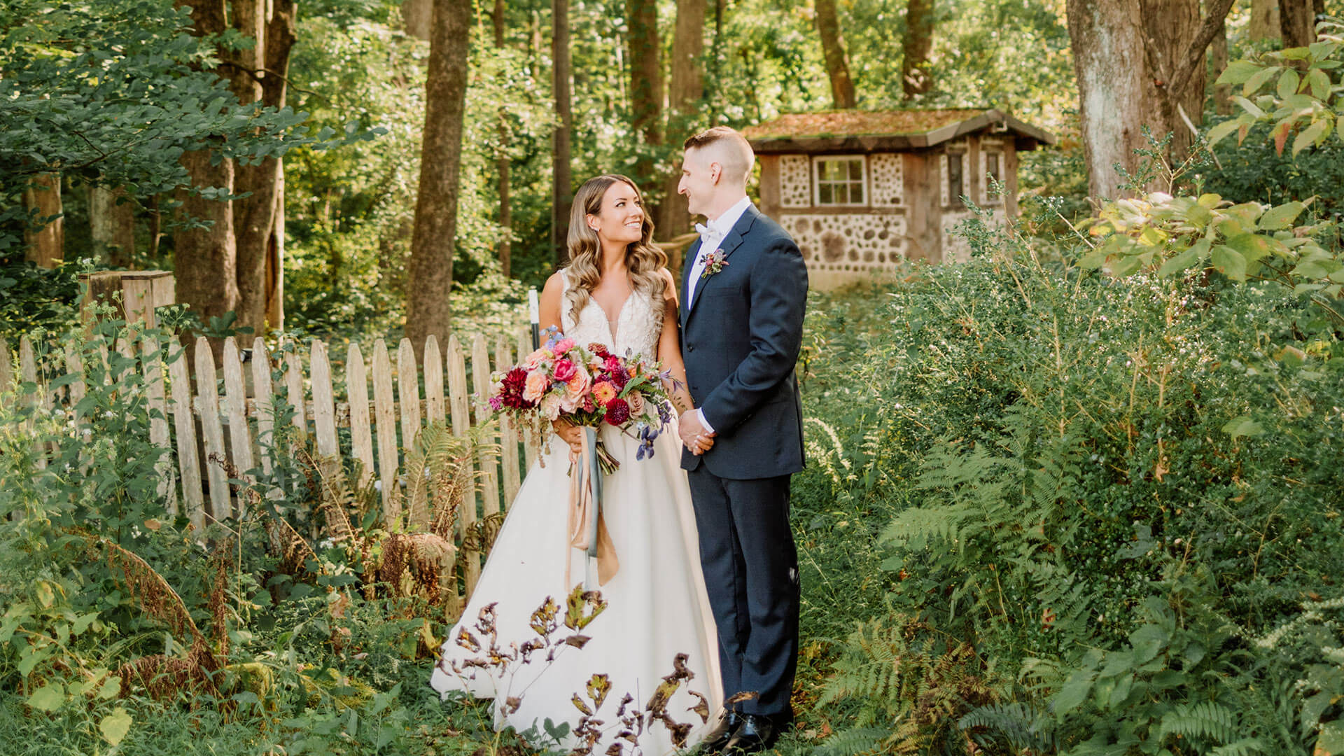 Bride and groom standing in a wooded area surrounded by foliage, looking at each other lovingly.