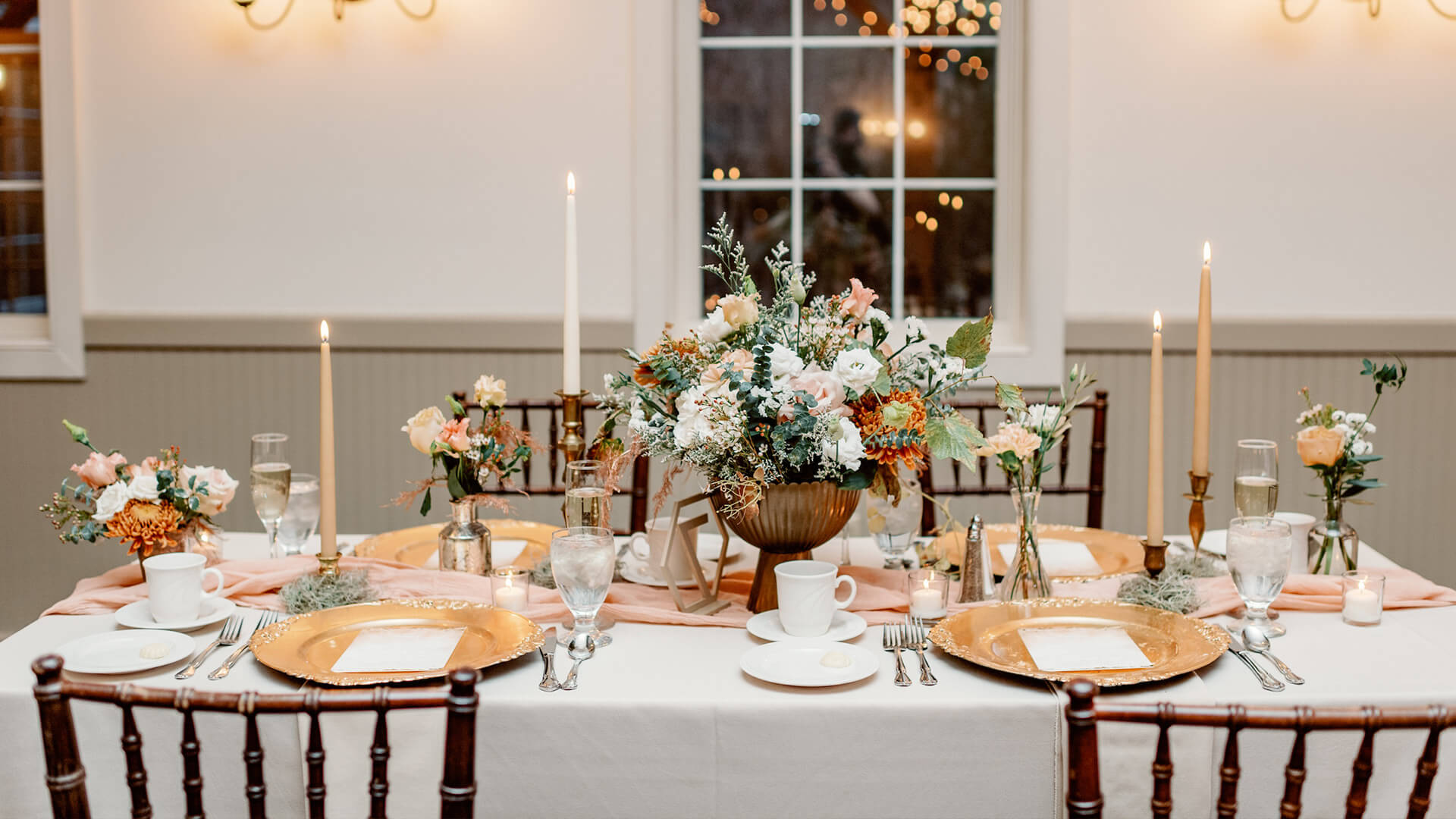 A white dining table adorned with candlesticks, flowers, glasses, silverware, and gold plates.